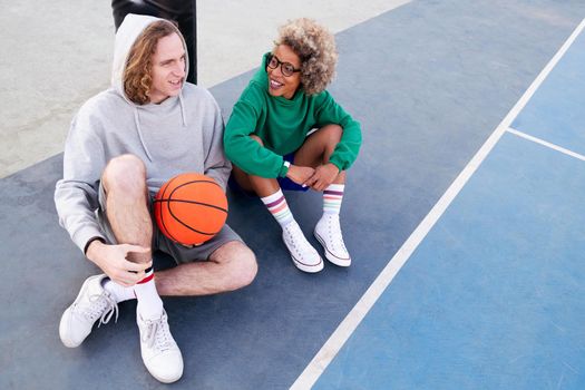 latin woman and caucasian man chatting happily sitting on the basketball court after the game, concept of friendship and urban sport in the street, copy space for text