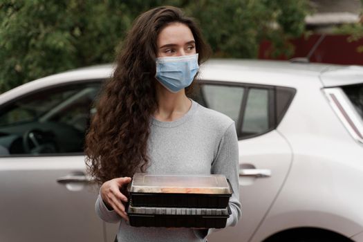 Sushi set in box healthy food delivery service by car. Attractive girl in medical mask with 2 sushi boxes stands in front of car