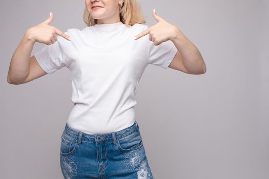 Cropped view from front of happy brunette standing in white shirt and jeans and pointing at outfit with fingers. Cheerful girl laughing and posing on grey isolated backround. Concept of casual style.