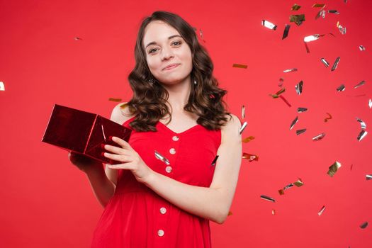 Portrait of adorable smiling young woman long curly hair posing isolated at red studio background. Beautiful happy female enjoying relaxing surrounded by shining colorful confetti looking at camera