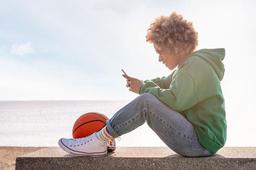 smiling young latin woman in a hoodie sitting on a bench by the beach with a basketball using her phone, concept of lifestyle and technology of communication, copy space for text