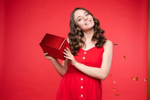 Portrait of adorable smiling young woman long curly hair posing isolated at red studio background. Beautiful happy female enjoying relaxing surrounded by shining colorful confetti looking at camera