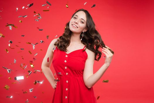 Studio portrait of beautiful young brunette lady with wavy hair in summer red dress with buttons smiling at camera. She is sprinkled or showered with sparkling confetti on red background. Isolate.