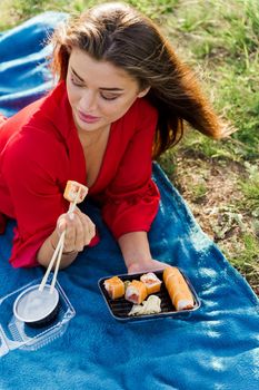 Attractive girl with 3 sushi sets lays on the blue cover on green hills background. Sexy girl in red dress gets sushi box from courier. Food delivery from restaurant