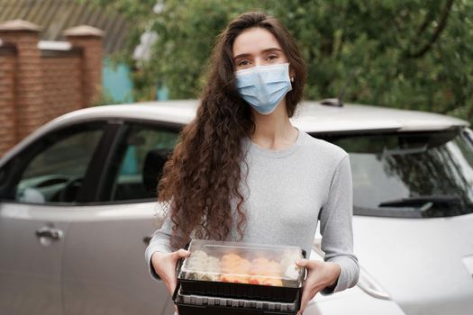 Sushi set in box healthy food delivery service by car. Girl courier in medical mask with 2 sushi boxes stands in front of car