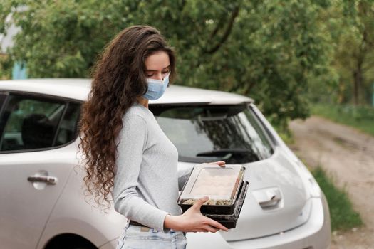 Sushi set in box healthy food delivery service by car. Girl courier in medical mask with 2 sushi boxes stands in front of car