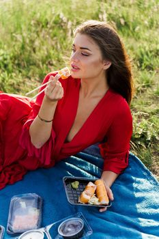 Sushi near lips of girl with blue eyes at picnic. Food delivery from japanese restaurant. Vertical photo advert for social networks. Pretty girl eats sushi set in the park.