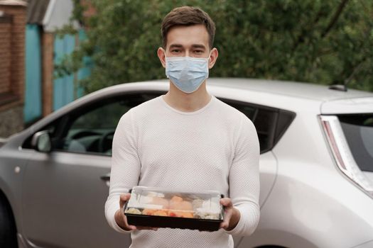 Sushi set in box healthy food delivery online service by car. Handsome man courier in medical mask gives sushi box to you. Japanese cuisine