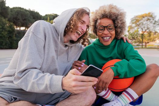 friends laughing while looking at things on the mobile phone sitting after playing basketball in a city park, concept of friendship and urban sport in the street