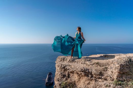 A girl with loose hair in a long mint dress descends the stairs between the yellow rocks overlooking the sea. A rock can be seen in the sea. Sunny path on the sea from the rising sun.