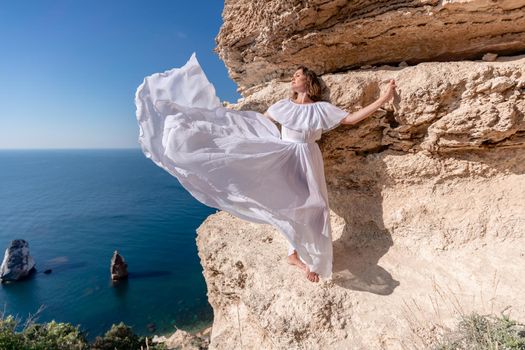 A beautiful young woman in a white light dress with long legs stands on the edge of a cliff above the sea waving a white long dress, against the background of the blue sky and the sea