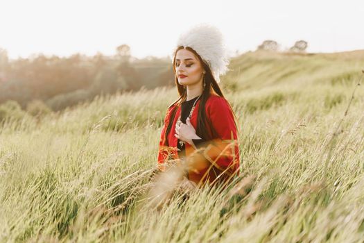 Georgian girl with closed eyes in white papakha and red national dress seats on the green grass and dreams. Georgian culture lifestyle.