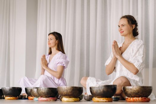 Two women are sitting with Tibetan bowls in the lotus position before a yoga class in the gym.
