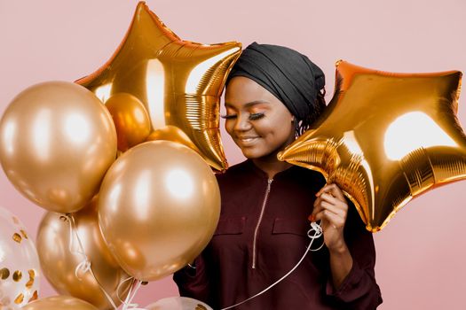 Muslim black girl with golden helium balloons smiles isolated pink background. Close-up african young woman celebrates her birthday