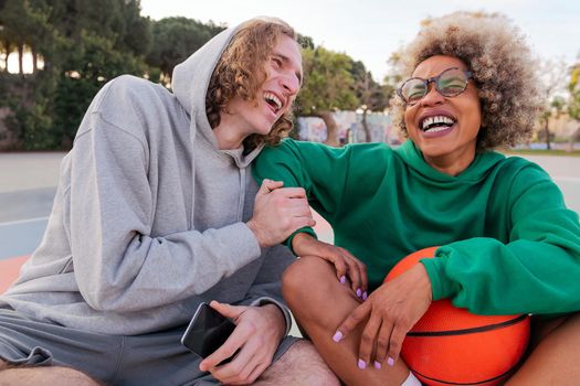 friends laughing while sharing a good time sitting after playing basketball in a city park, concept of friendship and urban sport in the street