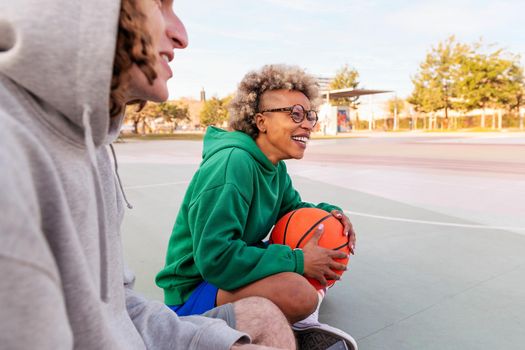 latin woman laughing as she has a good time sitting on the court with her friend after playing basketball in a city park, concept of friendship and urban sport in the street, copy space for text