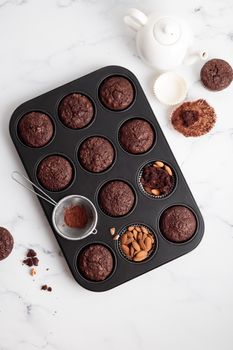 Freshly baked chocolate muffins in a baking pan on white table with ingredients