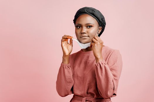 Muslim girl in medical mask on the pink background. Portrait of african woman who sick coronavirus covid-19