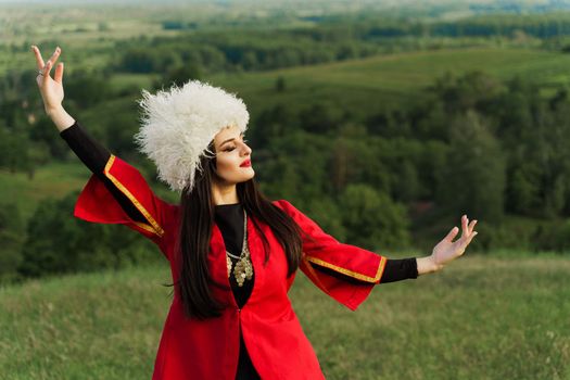 Georgian girl in white papakha dances national dance in red national dress on the green hills of Georgia background. Georgian culture lifestyle.