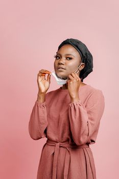 Muslim girl in medical mask on the pink background. Portrait of african woman who sick coronavirus covid-19. Black girl touches her mask. Vertical photo