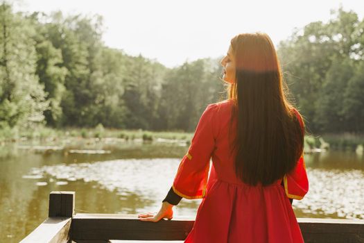 Georgian woman in red national dress with cross symbols. Attractive woman on the lake with forest background. Georgian culture lifestyle. Woman looks left side