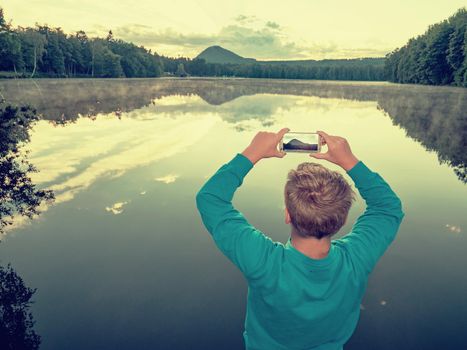 Blue sweatshirt guy is taking phone picture of pond and sunset. natural park with amazing lake and pure nature.