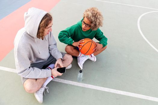 couple of friends chatting and sharing a good time sitting on the court after a basketball game in a city park, concept of friendship and urban sport in the street, copy space for text