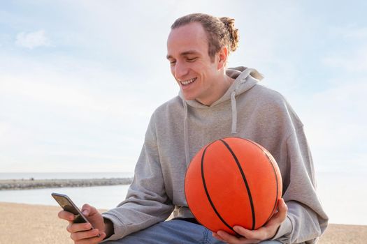 young caucasian man with a hoodie and a basketball smiling using his mobile phone, concept of lifestyle and technology of communication, copy space for text