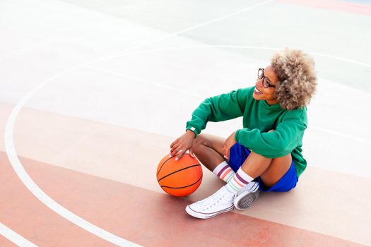 smiling latin woman resting sitting on the court after playing basketball, concept of urban sport outdoors, copy space for text