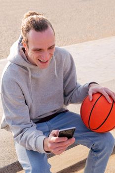 smiling caucasian man with a hoodie and a basketball using his mobile phone, concept of lifestyle and technology of communication
