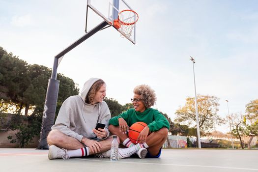 couple of friends laughing and having fun sitting on the court after a basketball practice at a city park, concept of friendship and urban sport in the street, copy space for text