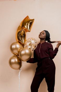 Black girl with golden ballons have a party and smile isolated on peach background. African woman celebrate graduation. Happy emotion of muslim young woman