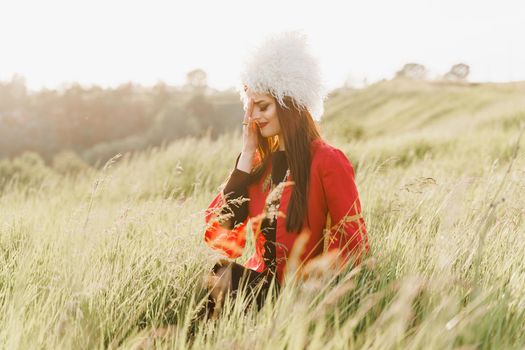 Georgian girl cry in white papakha and red national dress seats on the green grass and looks left side. Georgian culture lifestyle.