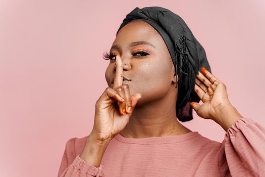 Muslim woman puts her finger to lips . Advert for discounts. African black attractive girl close-up portrait isolated on pink background
