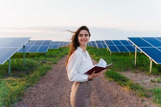 Investor woman stand and read book near solar panels row on the ground at sunset. Sustainability of planet