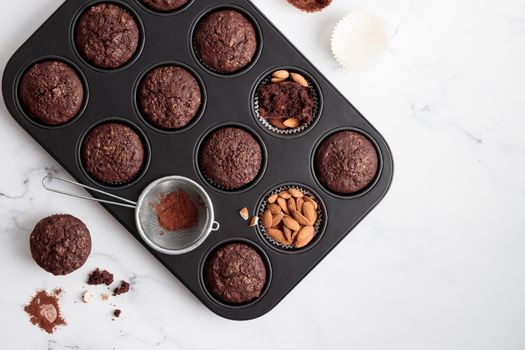 Freshly baked chocolate muffins in a baking pan on white table with ingredients