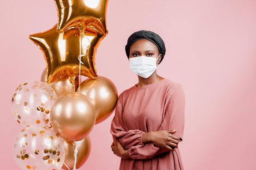 Muslim girl in medical mask on the pink background with golden baloons. Portrait close-up of african woman who sick coronavirus covid-19