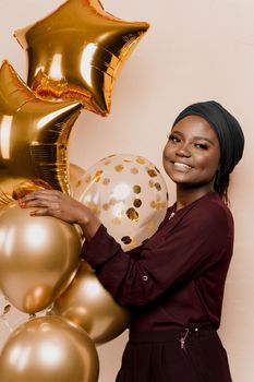 Muslim black girl with golden helium balloons isolated peach background. African young woman celebrates her birthday