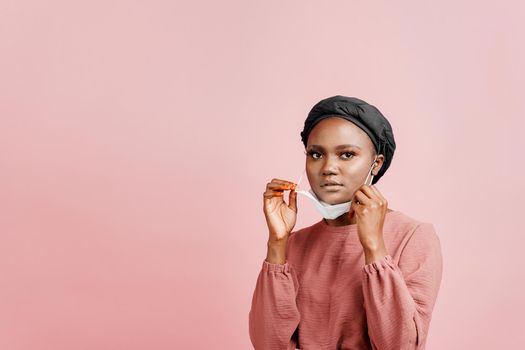 Muslim girl in medical mask on the pink background. Portrait of african woman who sick coronavirus covid-19. Black girl touches her mask.