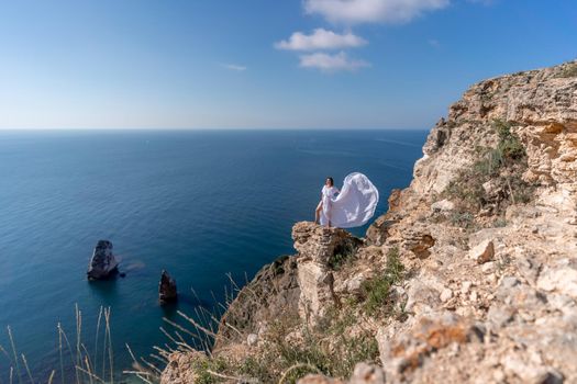 A beautiful young woman in a white light dress with long legs stands on the edge of a cliff above the sea waving a white long dress, against the background of the blue sky and the sea