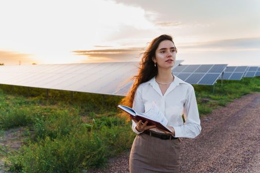 Investor woman stand and read book near solar panels row on the ground at sunset. Sustainability of planet