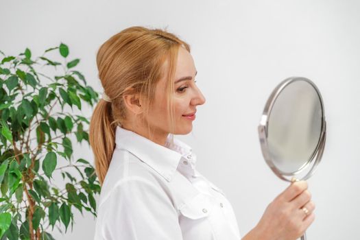 A blonde woman in white formal clothes looks at herself in the mirror. On a white background, green leaves at the back