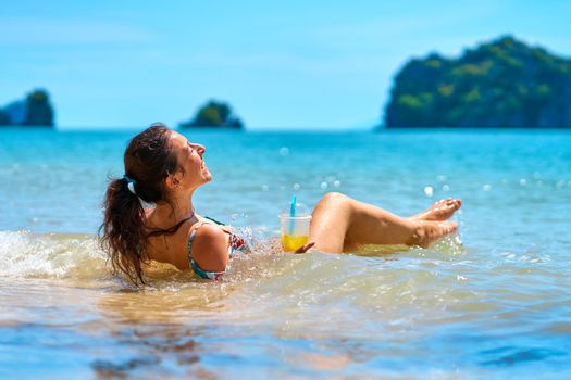 A beautiful slender girl in a swimsuit enjoys a tan while lying on a tropical beach, holding a glass of pineapple juice in her hand