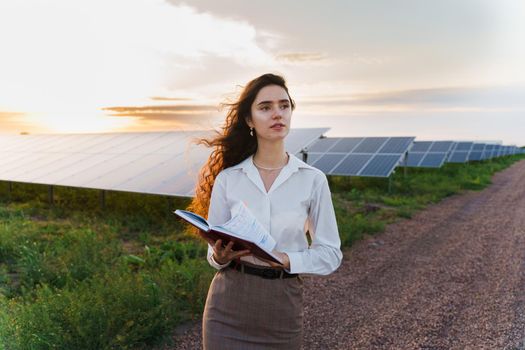 Investor woman stand and read book near solar panels row on the ground at sunset. Sustainability of planet