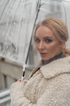 The blonde stands under a transparent umbrella during the rain. The fall season. Rear view. The woman is dressed in a black lace dress, her hair pulled back in a ponytail