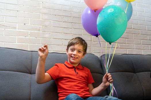 a happy European preschool boy is sitting on the sofa and smiling. The boy holds balloons inflated with helium by the ribbons. Birthday Celebration