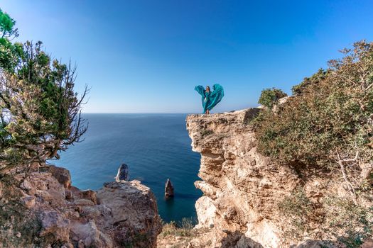 A girl with loose hair in a long mint dress descends the stairs between the yellow rocks overlooking the sea. A rock can be seen in the sea. Sunny path on the sea from the rising sun.