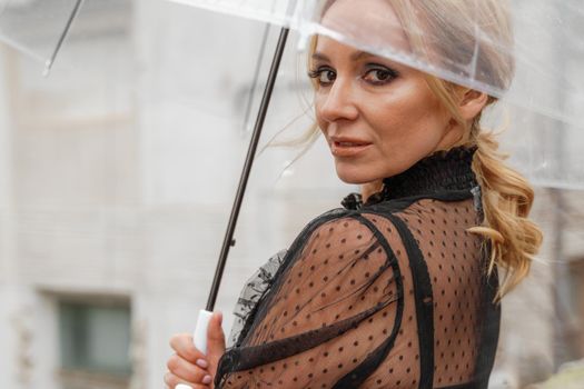 The blonde stands under a transparent umbrella during the rain. The fall season. Rear view. The woman is dressed in a black lace dress, her hair pulled back in a ponytail