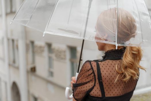 The blonde stands under a transparent umbrella during the rain. The fall season. Rear view. The woman is dressed in a black lace dress, her hair pulled back in a ponytail