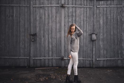 beautiful young blonde model girl. White pants. gray knitted sweater. black boots. wooden pendant on the neck in the form of a horse. On the Sunset. on a background of gray wooden gates